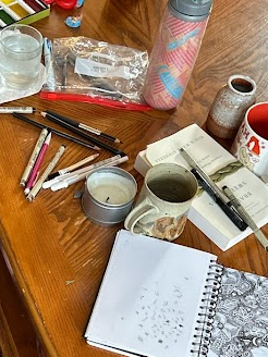 A wooden coffee table with a sketchbook, a novel, and some mugs, pens, a candle, and a water bottle. 